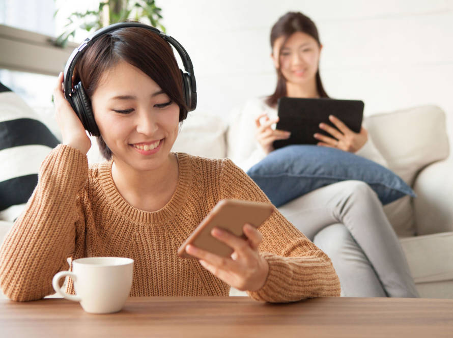 One female sitting in the front having headphones on and looking at her mobile phone while another female sitting at the back on a sofa looking at a tablet